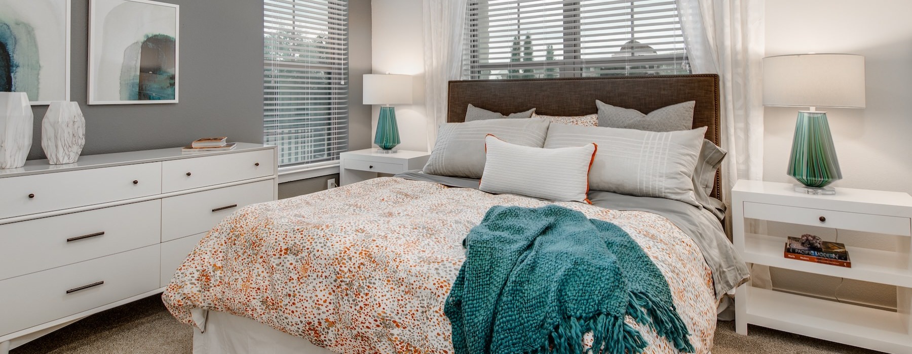 Model bedroom at Overture Stone Oak apartments in San Antonio, TX, featuring a bed, bedside table, dresser, and windows.