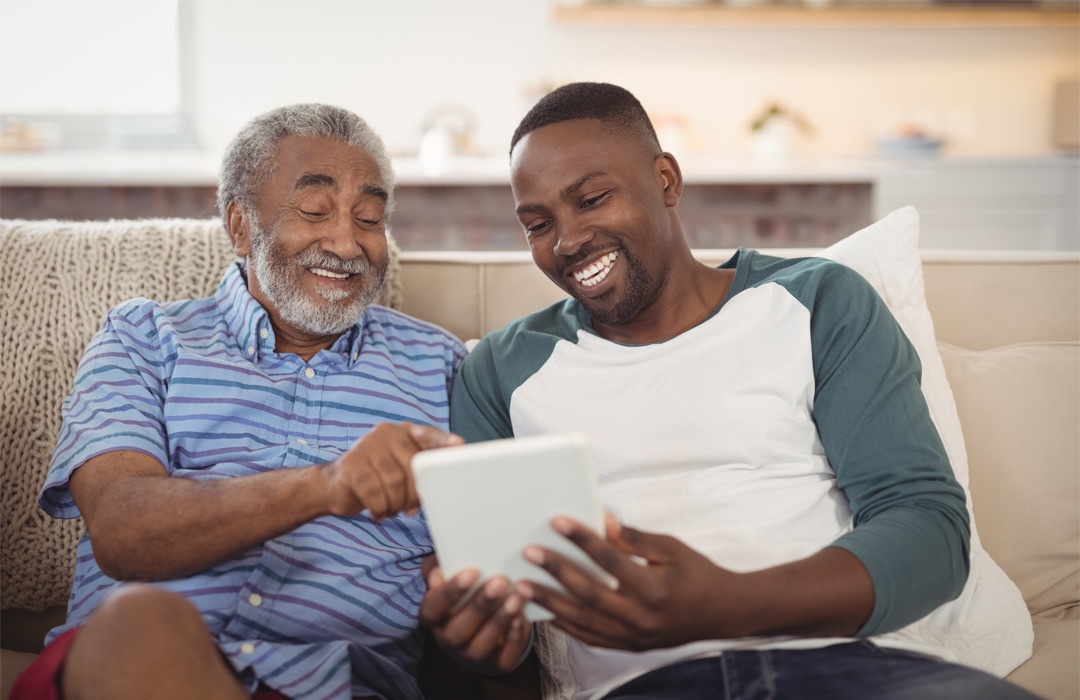 Understanding Your Choices Father and son sitting on a couch in a living room looking at a tablet together.